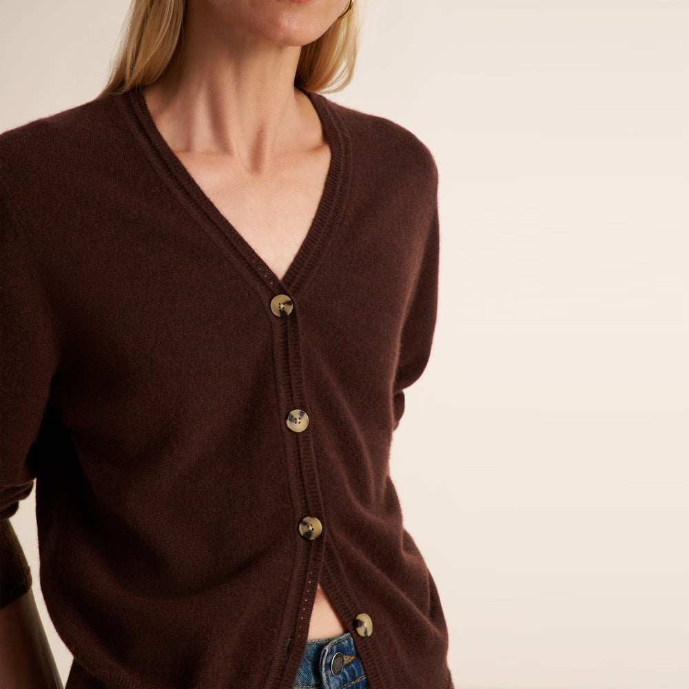 A close-up of a woman wearing a dark brown V-neck cardigan with gold buttons, paired with blue jeans, against a plain light beige background.