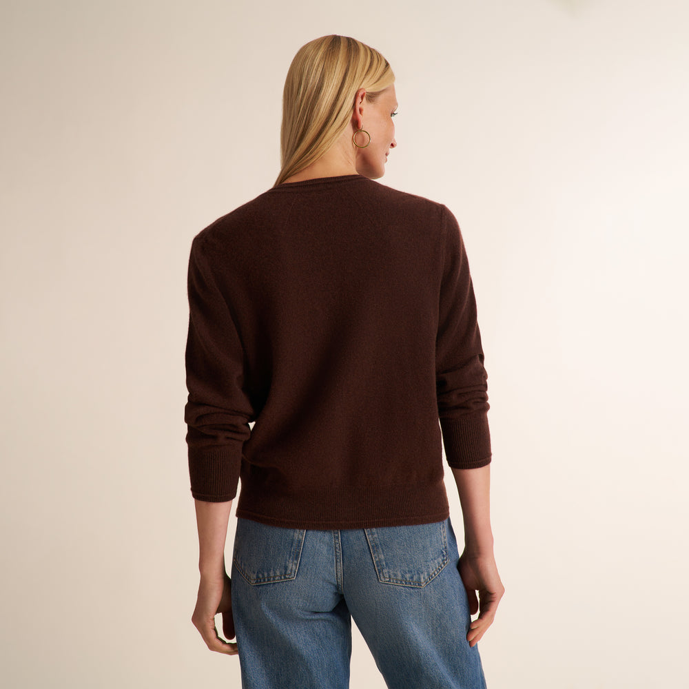 A woman viewed from the back wearing a dark brown crewneck sweater and blue jeans, standing against a plain light beige studio background.