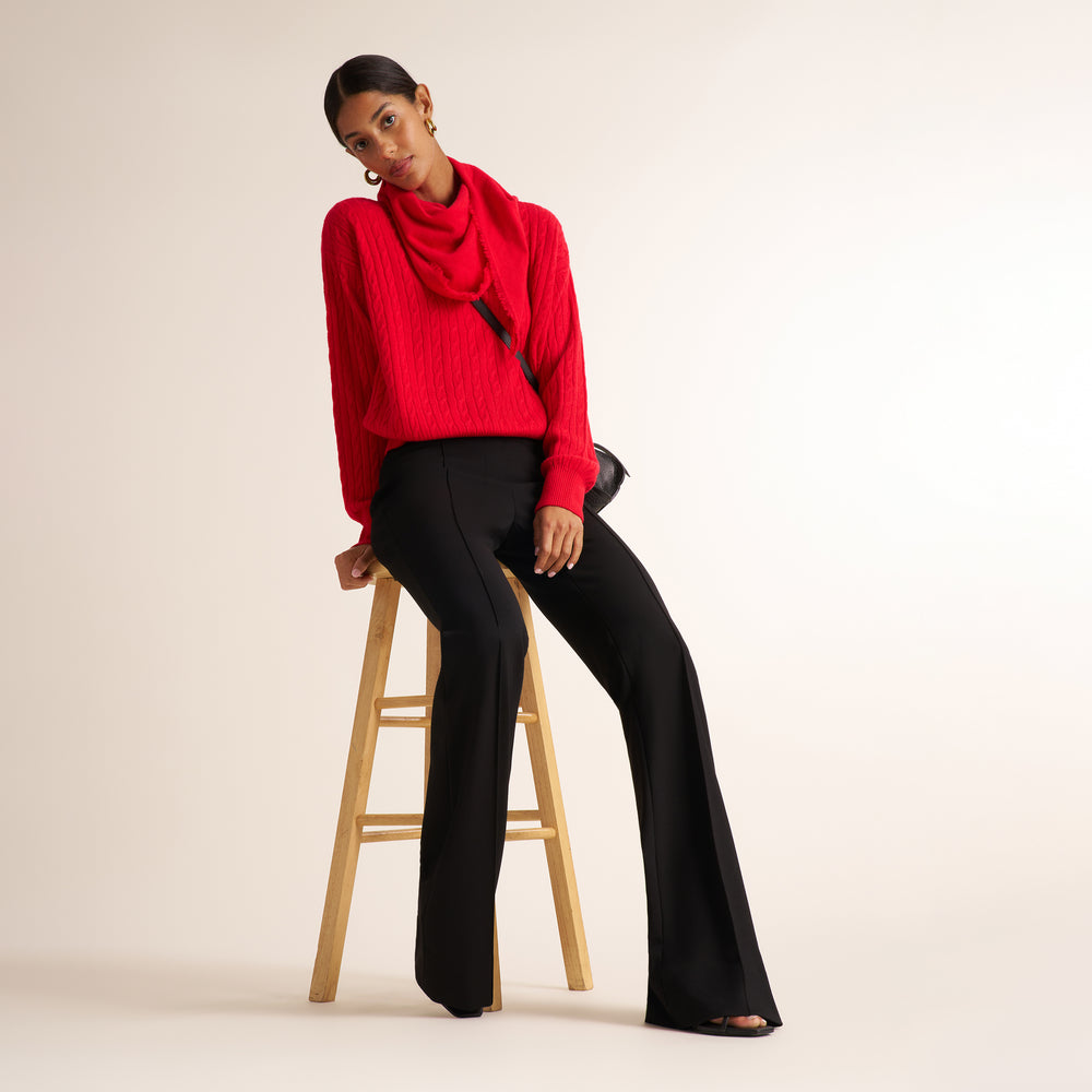 A model wearing a bright red cable-knit cowl-neck sweater and black wide-leg trousers, sitting on a wooden stool against a warm off-white studio background.