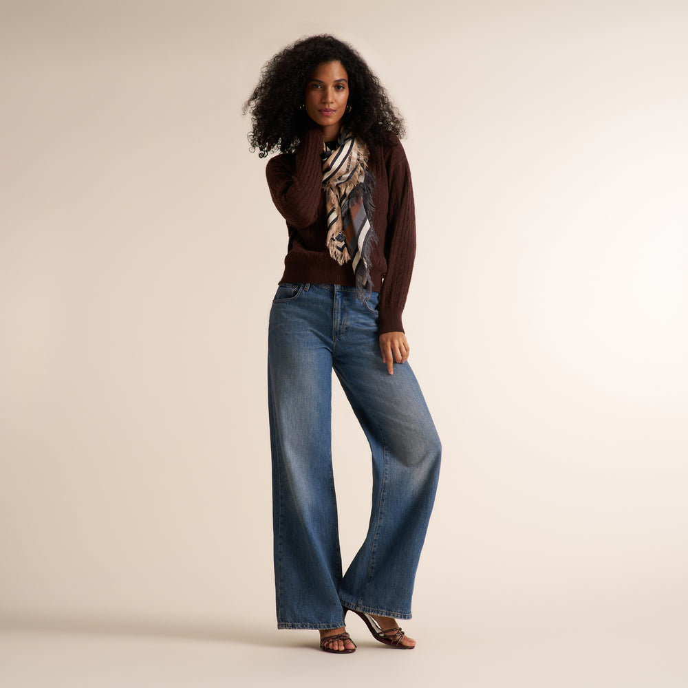 A model wearing a dark brown ribbed knit sweater, wide-leg blue jeans, a patterned scarf, and brown heeled sandals, standing against a solid light beige studio background.