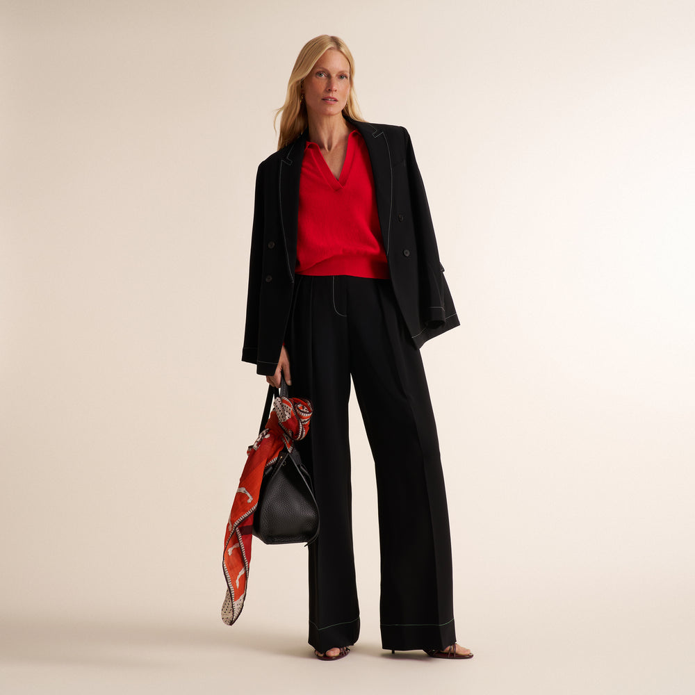 A model stands against a warm beige studio background wearing a black double-breasted blazer, wide-leg trousers, and a bright red V-neck top. She holds a black leather handbag with a red and orange patterned scarf draped over it.