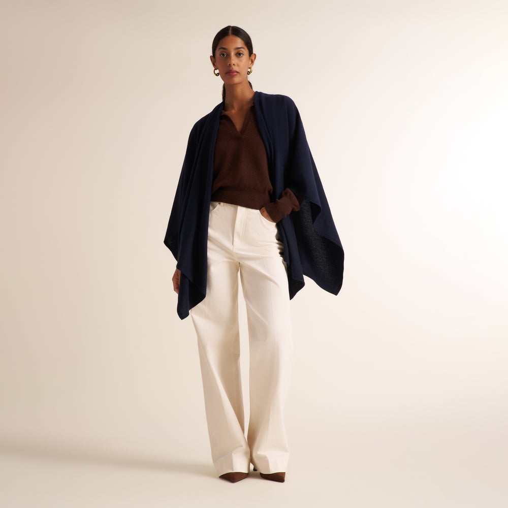 A model stands against a warm beige studio background wearing a dark navy draped wrap or poncho over a brown collared top, paired with high-waisted off-white wide-leg trousers.