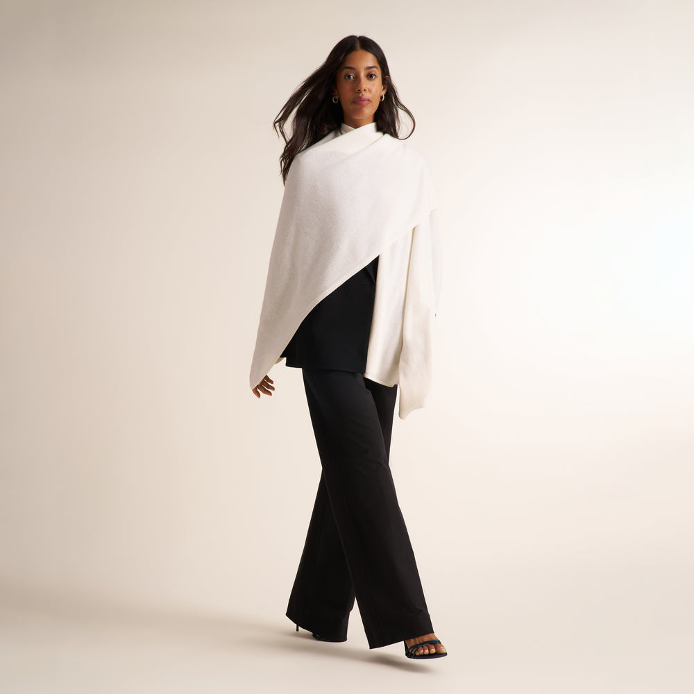 A woman modeling a white draped wrap or poncho-style top over a black outfit, standing against a plain warm off-white studio background.