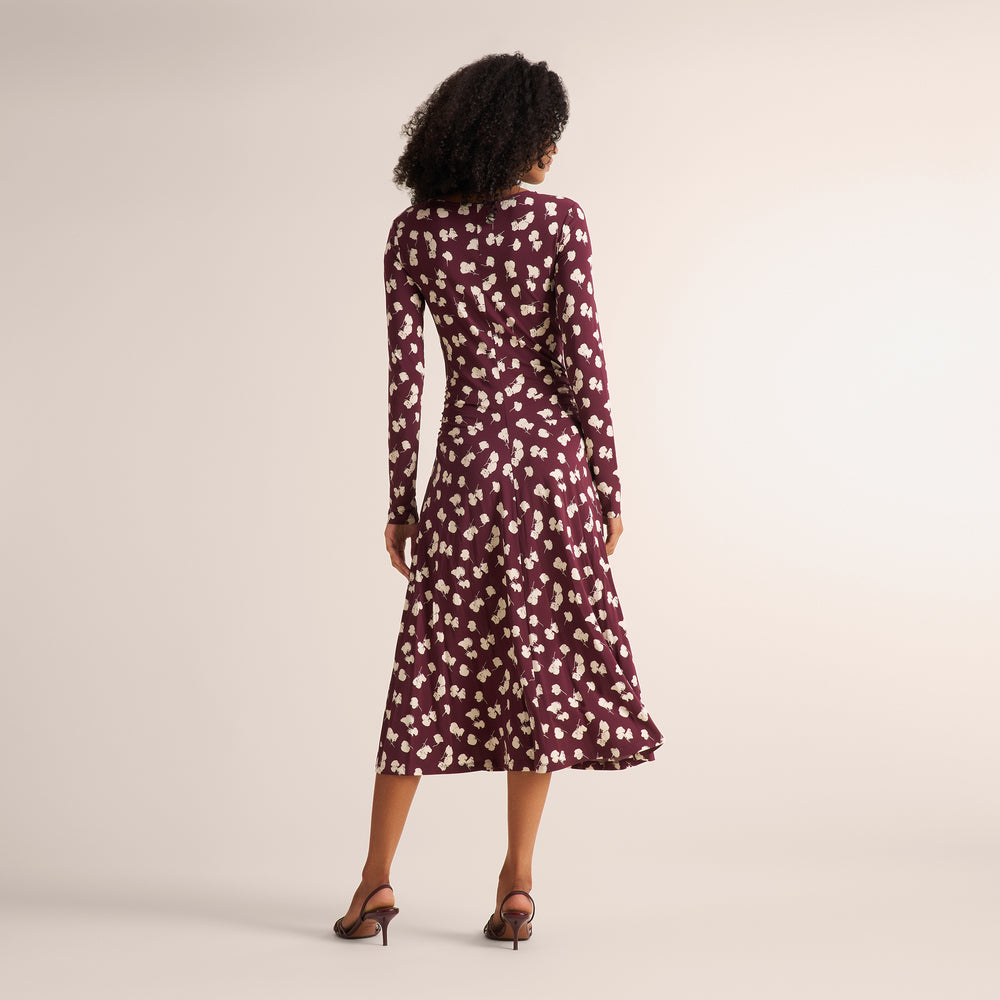 A woman viewed from behind wearing a long-sleeve midi dress with a burgundy base and a white floral pattern, paired with brown strappy heels, against a plain beige studio background.
