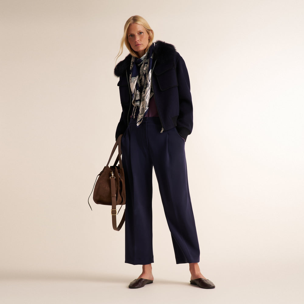 A woman models a dark navy blue puffer jacket with a fluffy collar, paired with matching wide-leg trousers, a patterned scarf, and a brown leather handbag, set against a plain beige studio background.