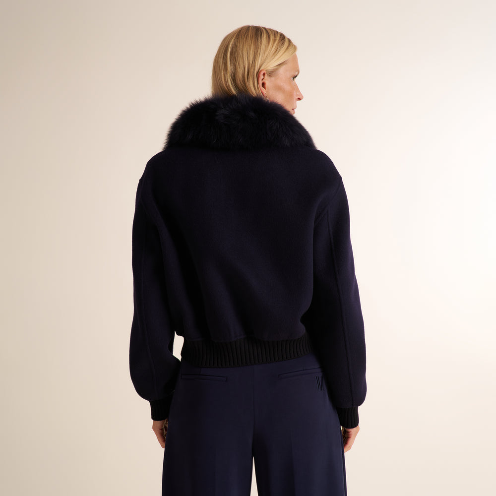 A woman shown from the back wearing a dark navy knit sweater with a large black fur collar and matching dark navy trousers, against a plain light beige studio background.