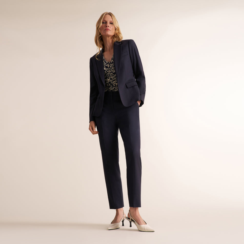 A woman models a dark navy blue two-piece pantsuit, consisting of a single-breasted blazer and matching trousers, paired with a patterned top and white pointed-toe heels, set against a plain beige studio background.