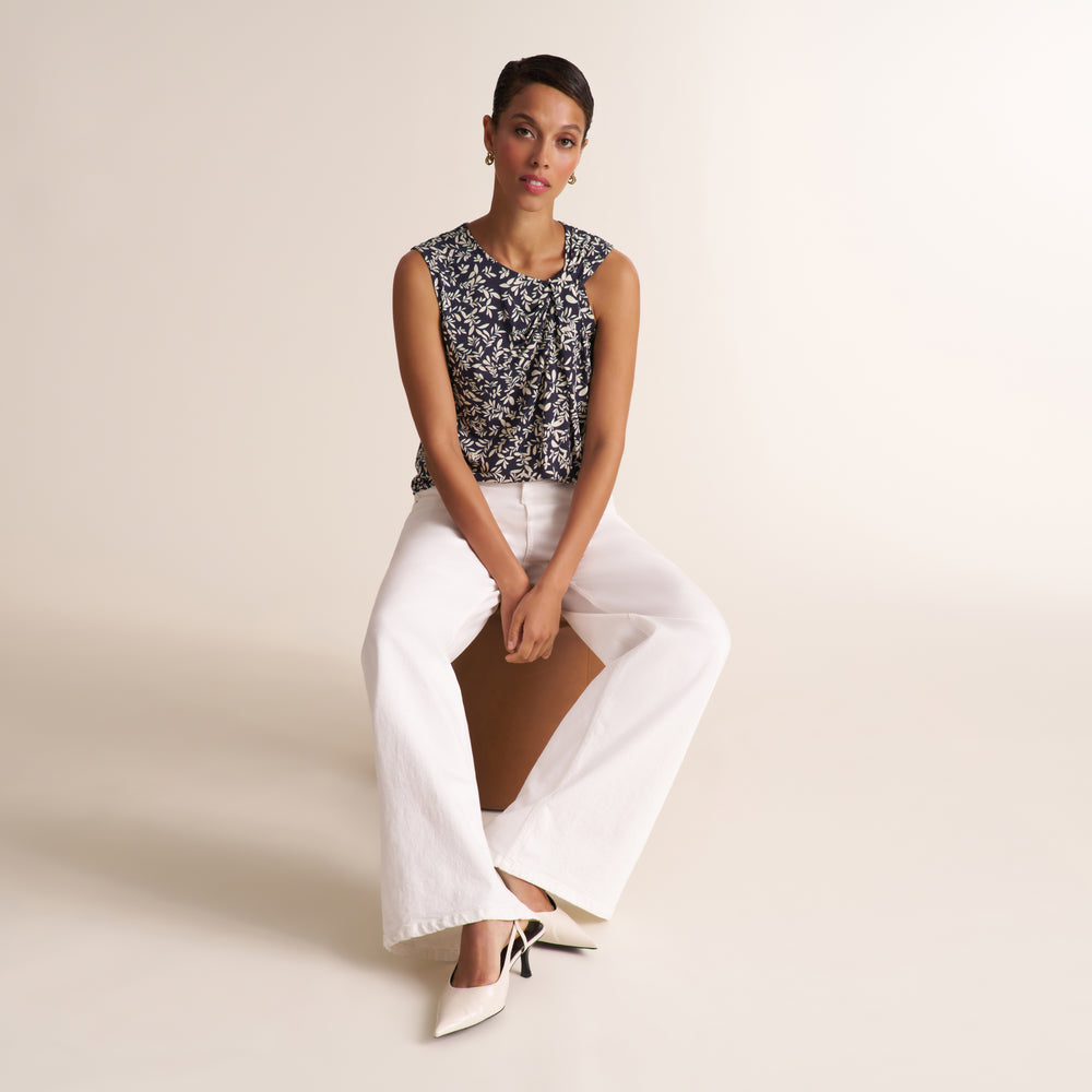 A woman seated on a studio floor wearing a sleeveless black-and-white floral top, white wide-leg pants, and white pointed heels against a plain warm beige background.