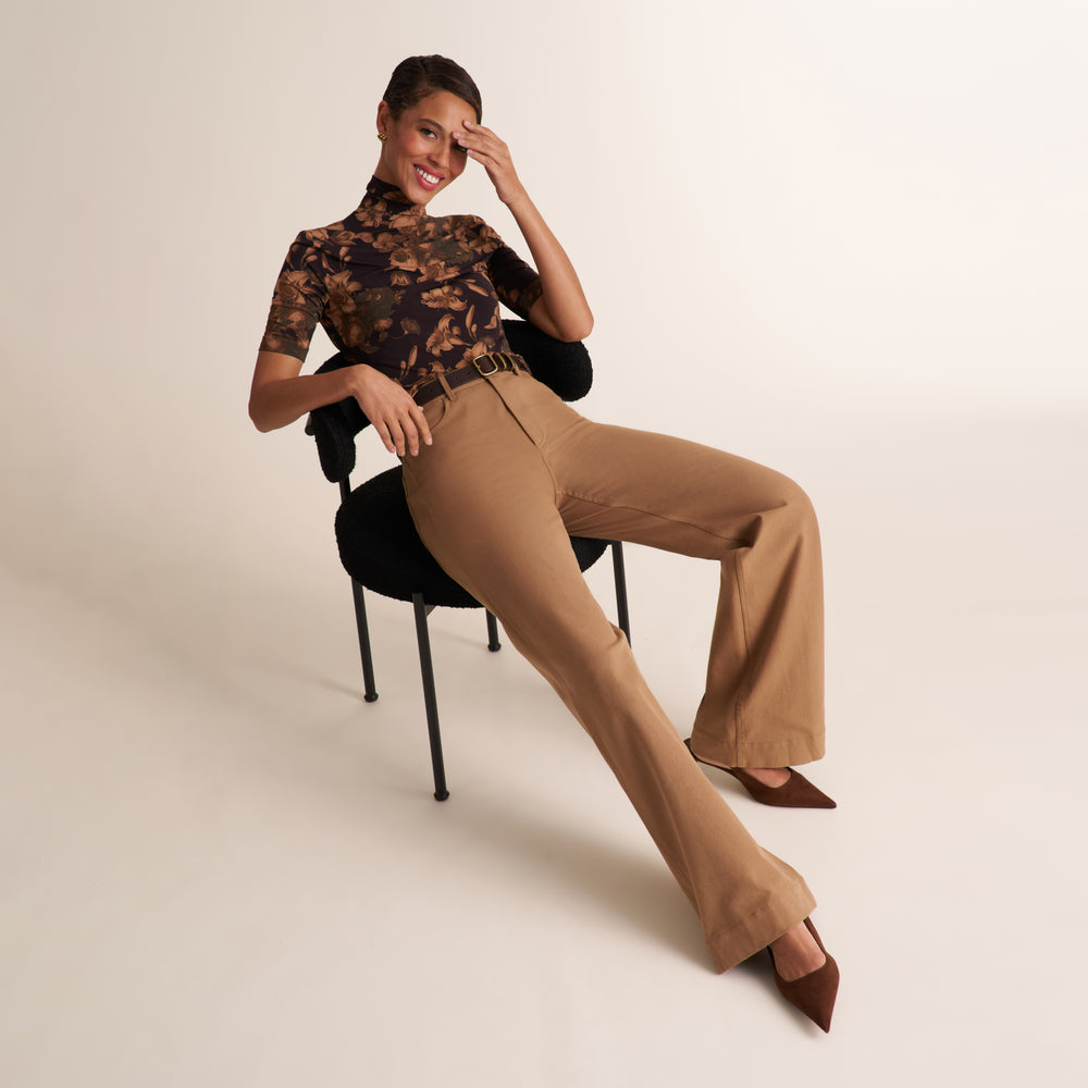 A woman poses on a black chair wearing tan wide-leg trousers, a dark floral short-sleeve top, and brown pointed-toe heels against a plain light beige studio background.