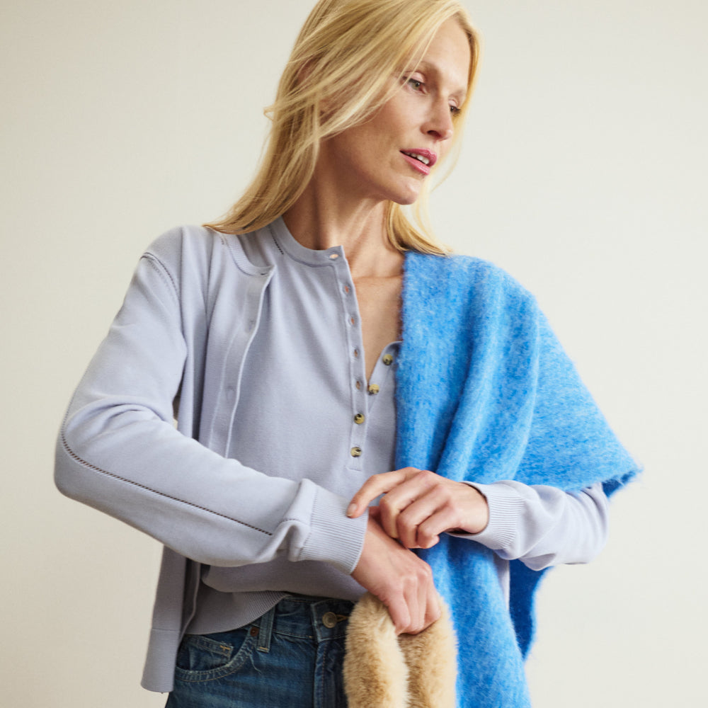 Woman wearing a light blue cardigan over a gray top, holding a beige handbag against a plain background.