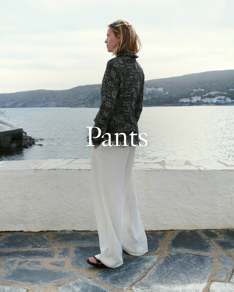 Woman wearing white pants and a black patterned top by a waterfront. "Pants" overlaid on the image