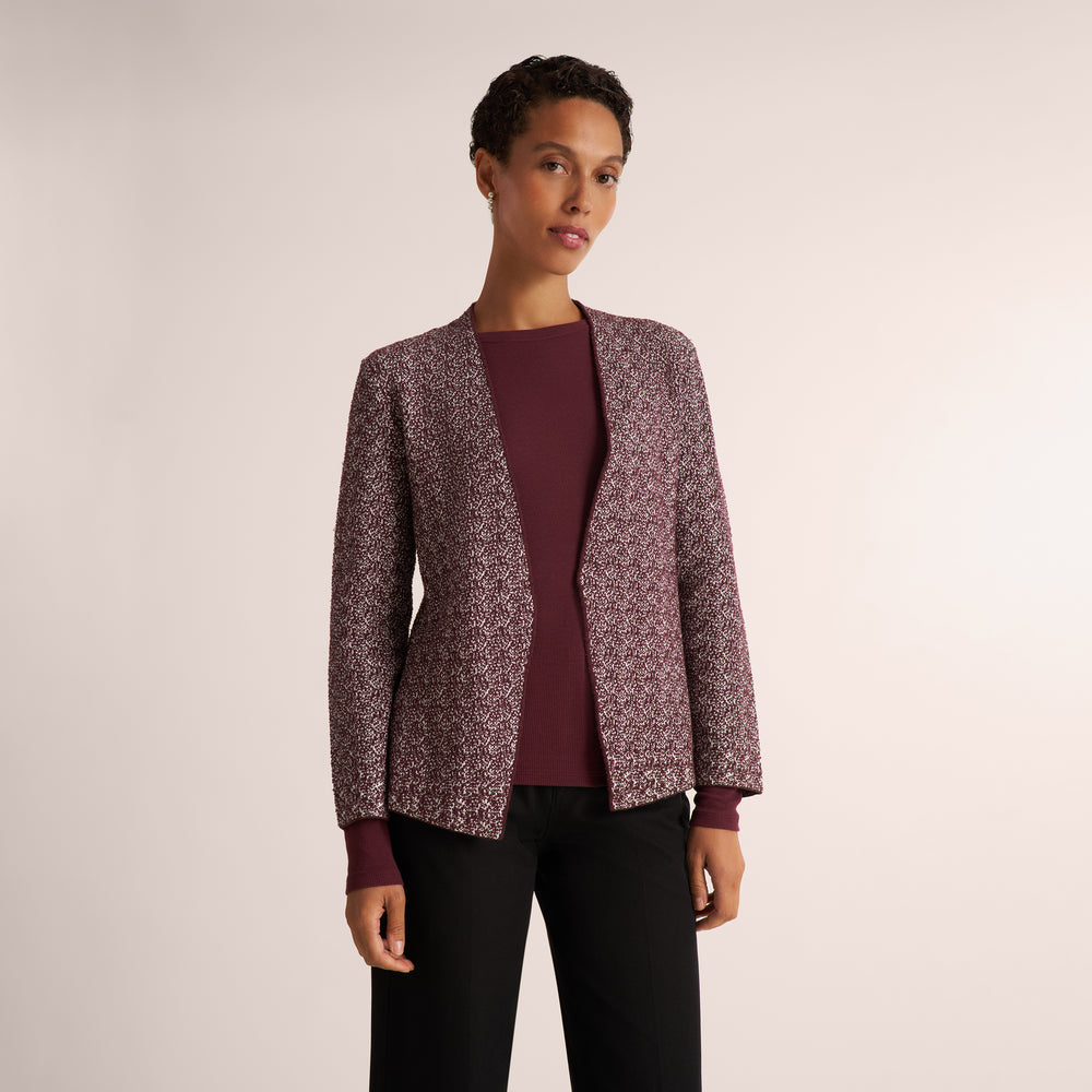 A model wearing a textured tweed blazer in a dusty rose and white pattern over a burgundy top and black pants, shot against a plain light beige studio background.