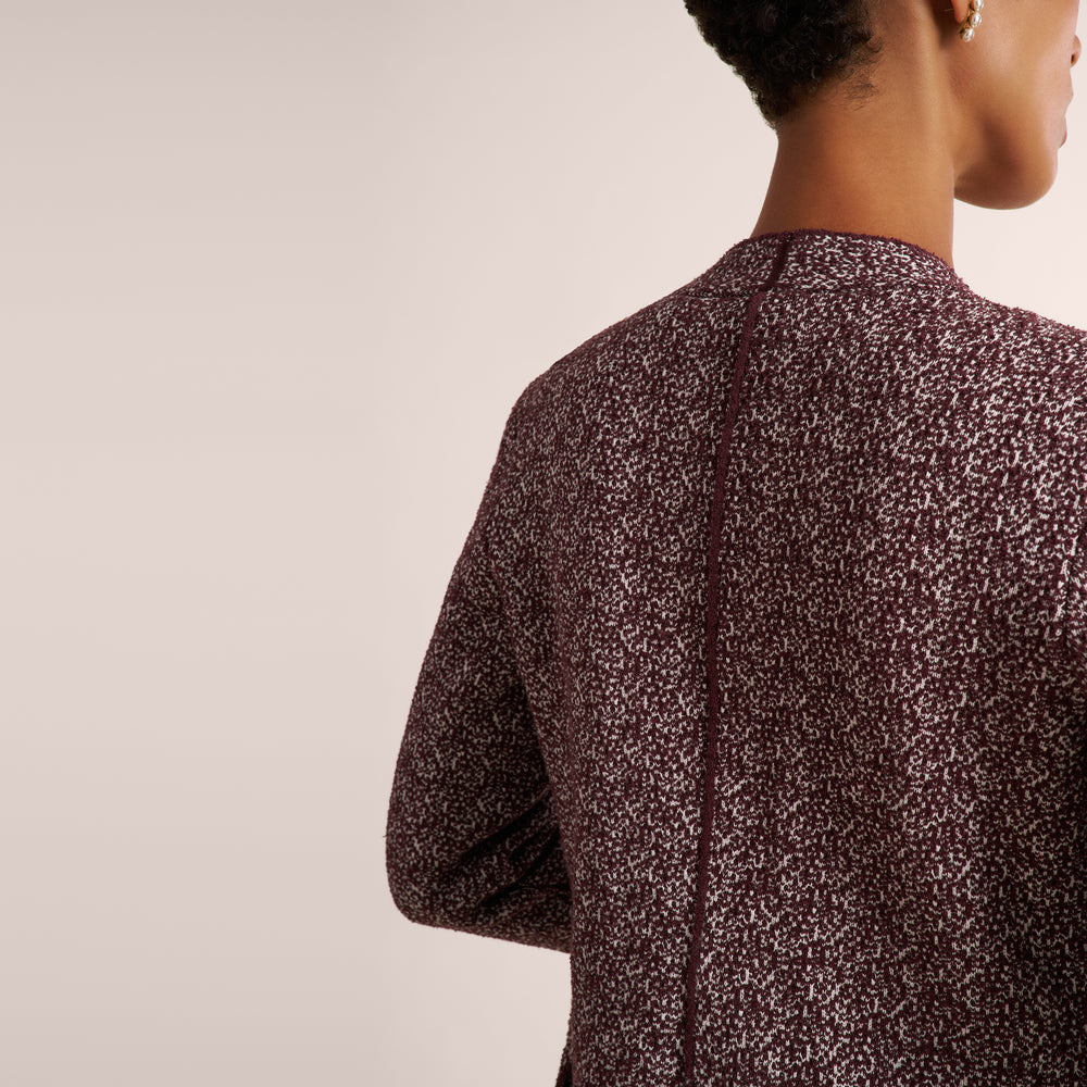 A close-up back view of a person wearing a long-sleeve top with a dense small-scale abstract print in dark burgundy and off-white, set against a plain light beige studio background.