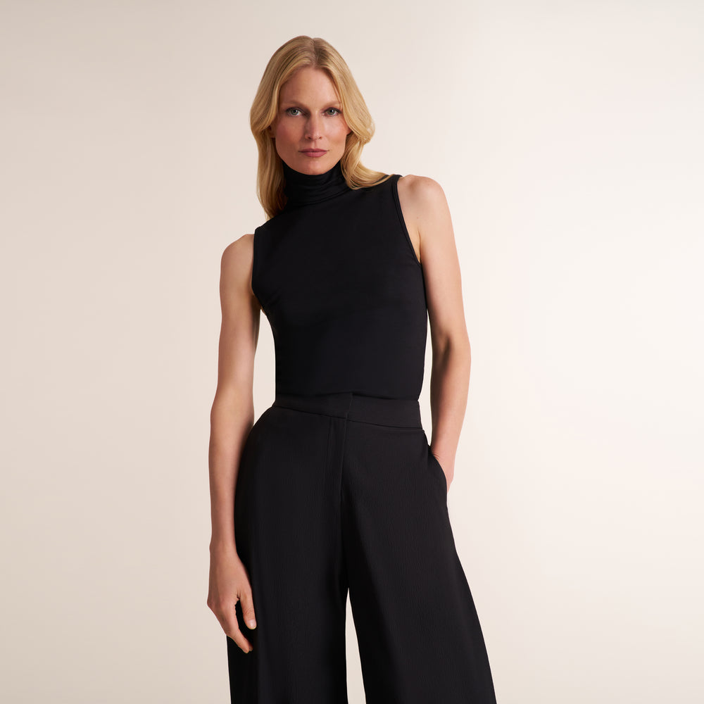 A blonde model wearing a black sleeveless mock-neck top and matching black wide-leg trousers, standing against a solid light beige studio background.