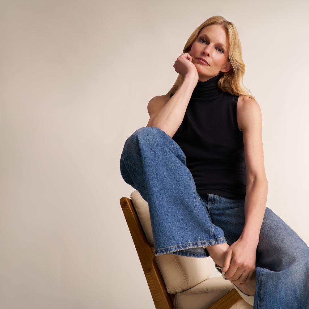 A woman with blonde hair sits on a wooden chair with a cream cushion, wearing a black sleeveless mock-neck top and wide-leg blue jeans against a plain beige background.