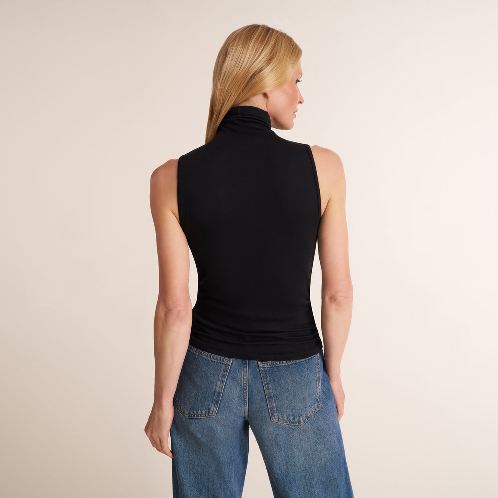 A woman shown from the back wearing a black sleeveless mock-neck top and blue jeans, standing against a plain light beige studio background.