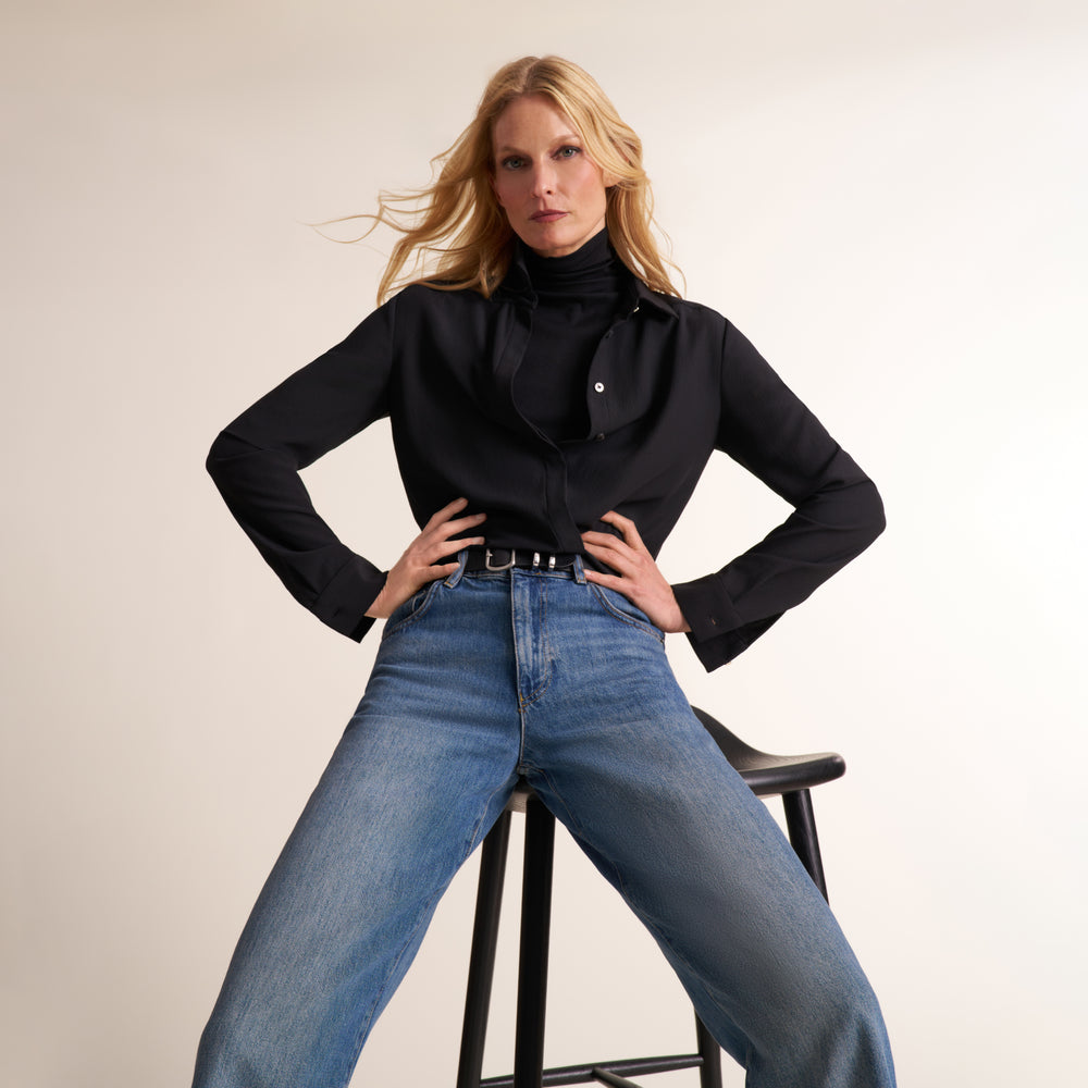 A blonde woman poses on a black stool wearing a black long-sleeve turtleneck blouse and high-waisted blue jeans against a plain light beige studio background.