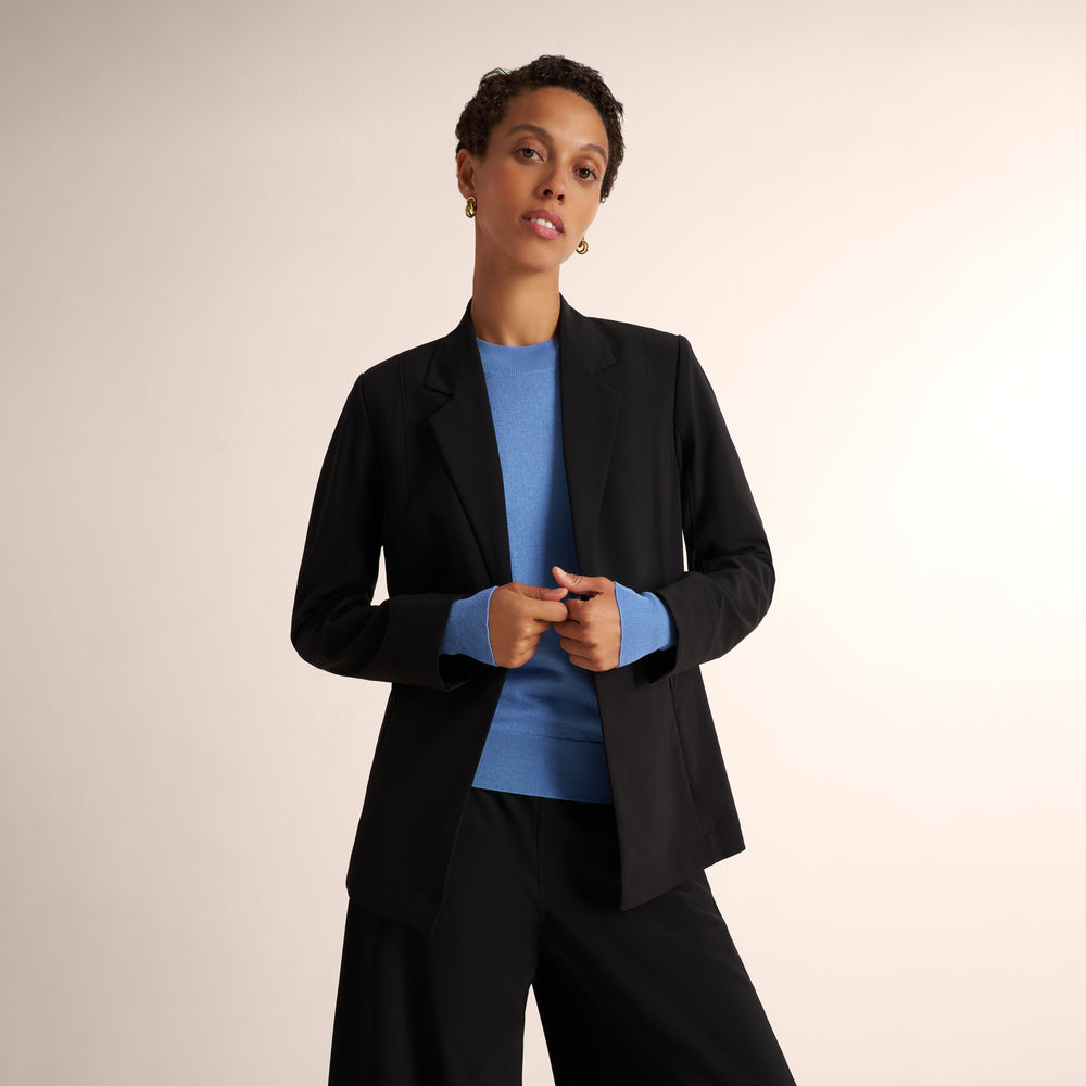 Woman wearing a black blazer and matching wide leg black pants with a blue splendor sweater underneath standing on a beige background