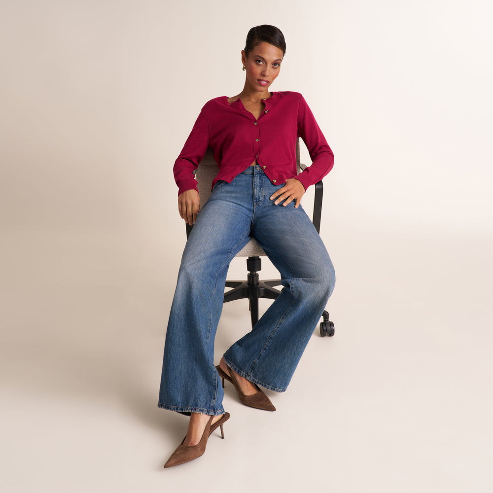A model sits on a black office chair wearing a cropped magenta button-up shirt, wide-leg blue jeans, and brown pointed-toe heels against a plain beige studio background.