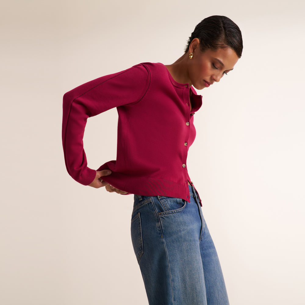 A woman wearing a bright magenta long-sleeve knit cardigan with buttons and blue jeans, posing against a plain beige studio background.