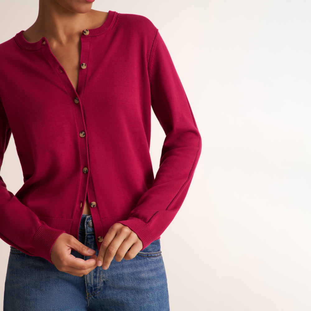 A person wearing a long-sleeve button-front cardigan in a rich magenta-pink color, paired with blue jeans, against a plain off-white background.