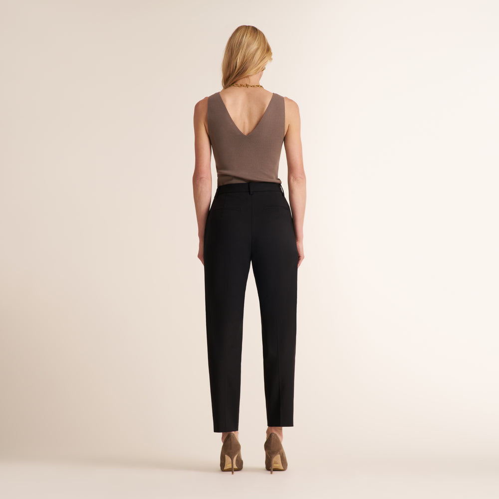 A woman shown from the back wearing a sleeveless taupe-brown top with a deep V-neck and black trousers, standing against a plain light beige studio background.
