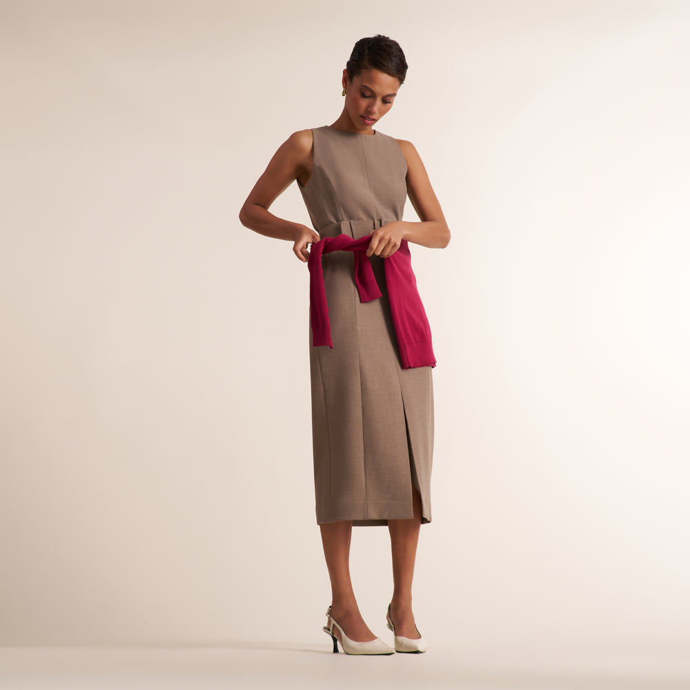 A woman modeling a sleeveless taupe midi dress with a side slit, tied at the waist with a bright magenta wrap or scarf, against a plain beige studio background.