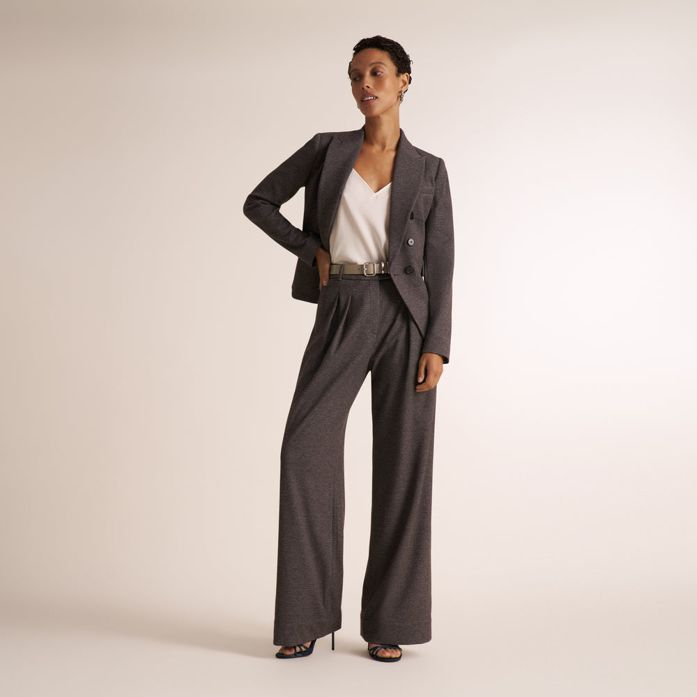 A woman on a light beige studio floor wearing a white spaghetti-strap camisole top and matching gray melange suit of a double breasted blazer and wide leg pants