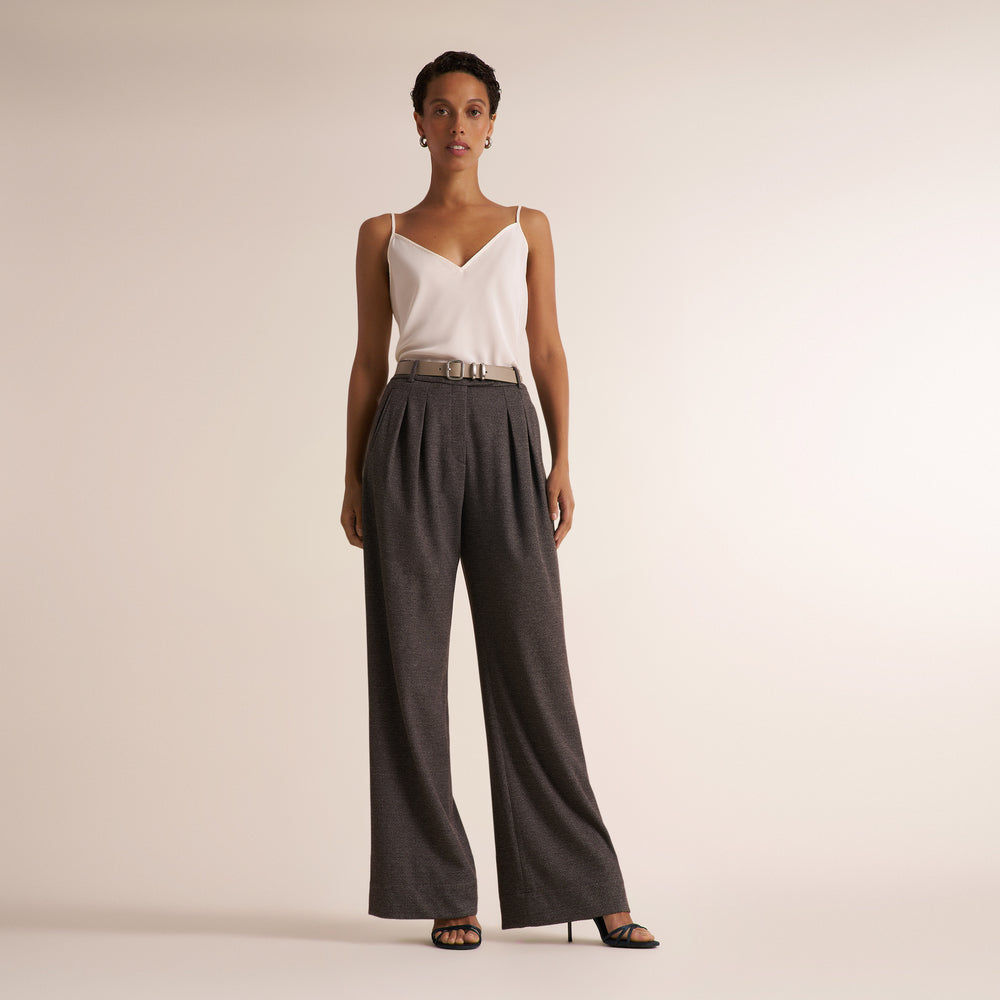 A woman on a light beige studio floor wearing a white spaghetti-strap camisole top and matching gray melange suit of a double breasted blazer and wide leg pants