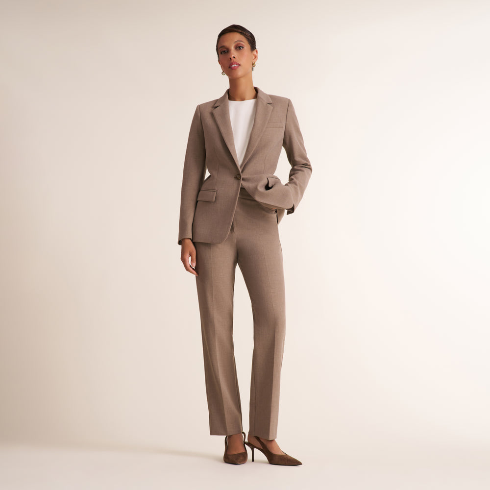 A model wearing a tailored taupe-brown two-piece suit with a blazer and matching trousers, styled over a white top and brown heels, standing against a plain light beige studio background.
