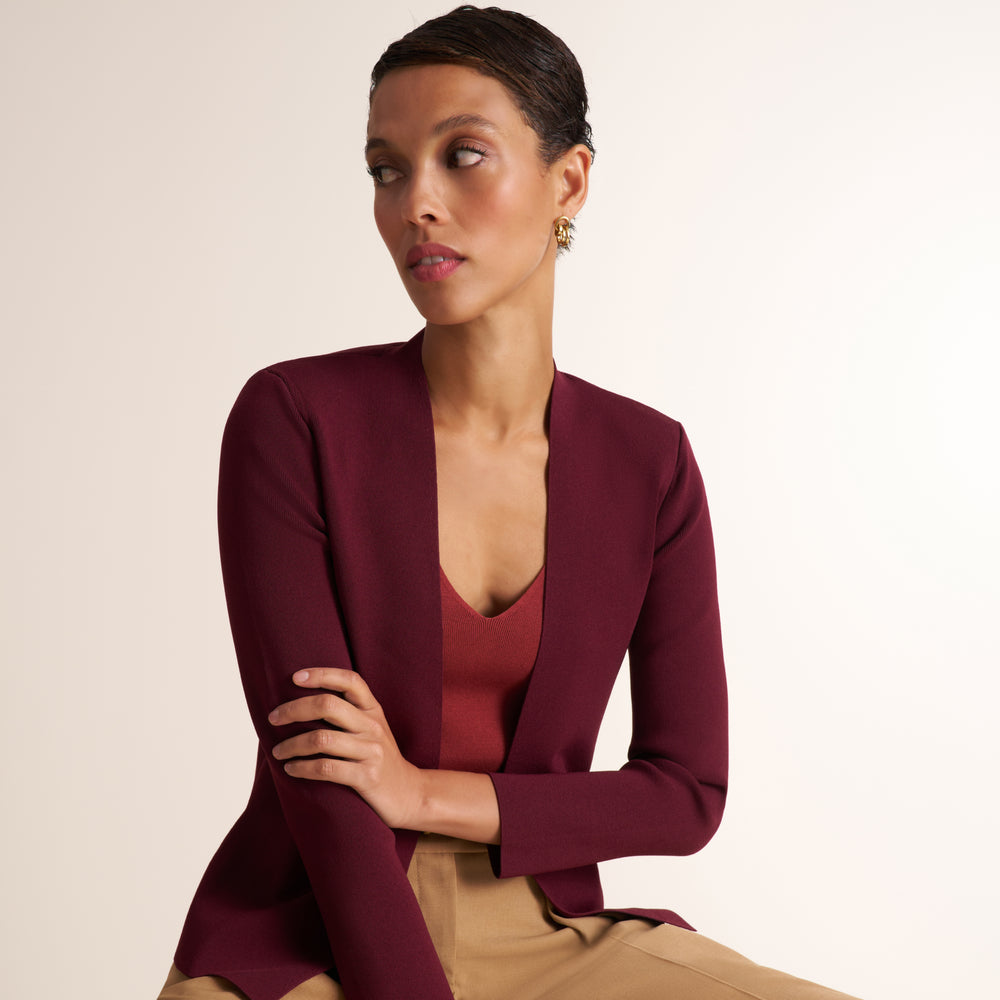 A woman wearing a deep burgundy open-front blazer over a matching burgundy top and tan pants, posed against a clean off-white studio background.