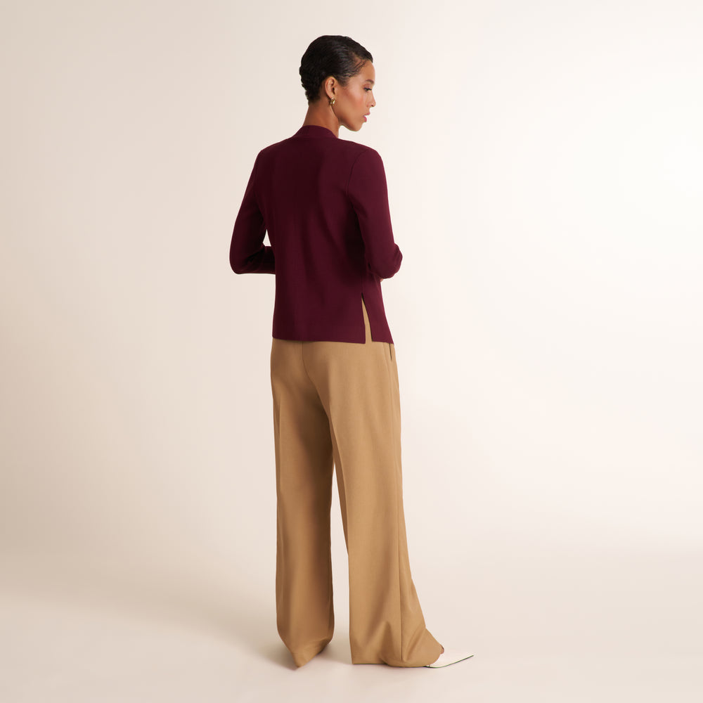 A woman is shown from the back wearing a dark burgundy long-sleeve top and beige wide-leg trousers, standing against a plain light beige studio background.