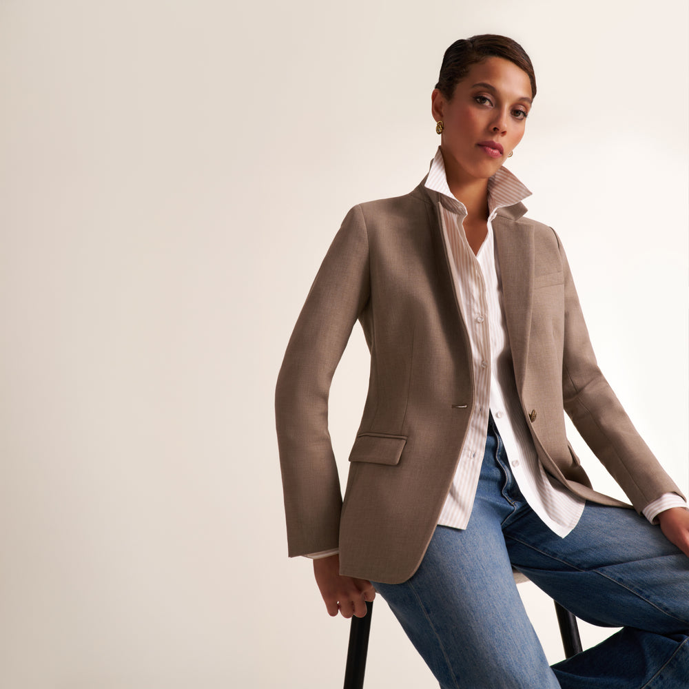 A woman models a taupe blazer over a white striped button-down shirt and blue jeans, seated on a stool against a plain off-white studio background.