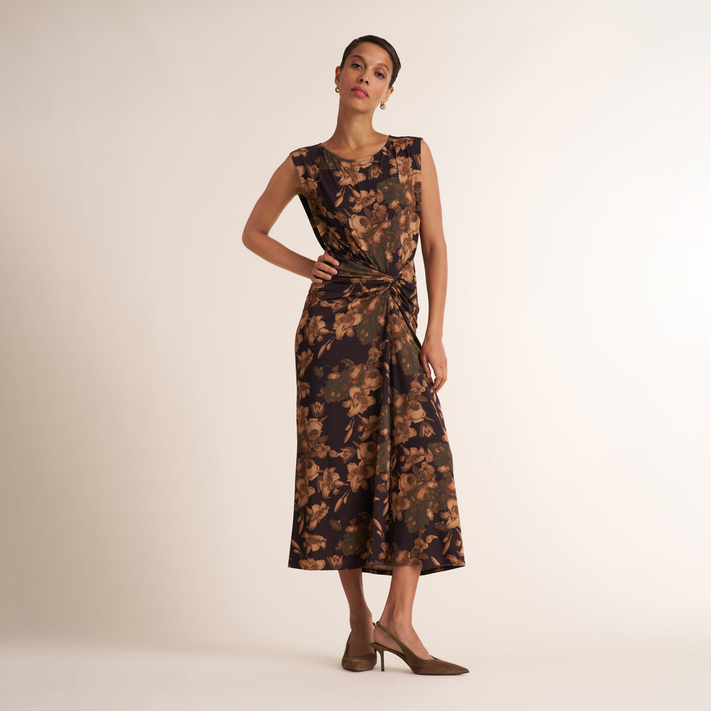 A woman models a sleeveless midi dress with a dark brown and black floral pattern and a twisted waist detail, paired with brown pointed-toe heels against a plain beige studio background.
