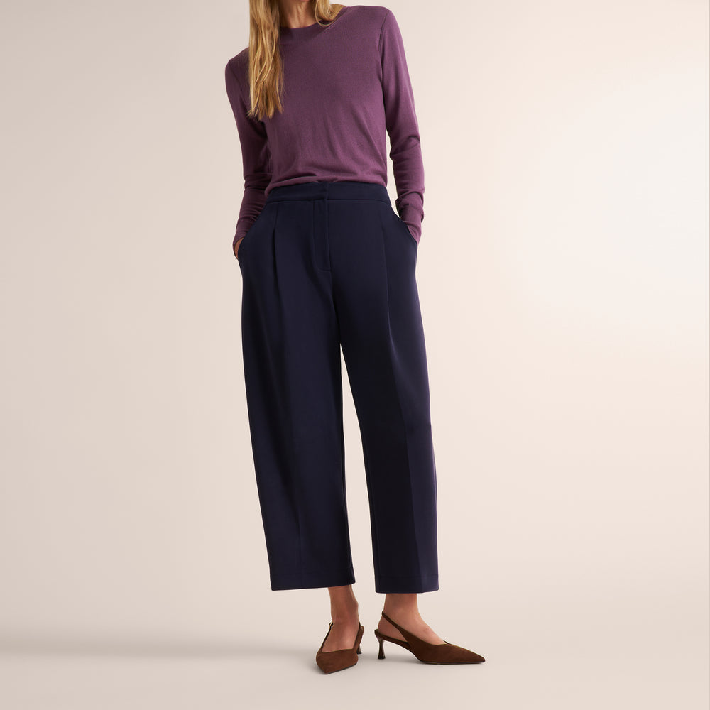 A model is shown from the waist down wearing navy blue wide-leg cropped trousers, a purple long-sleeve top, and brown slingback heels against a solid light beige background.