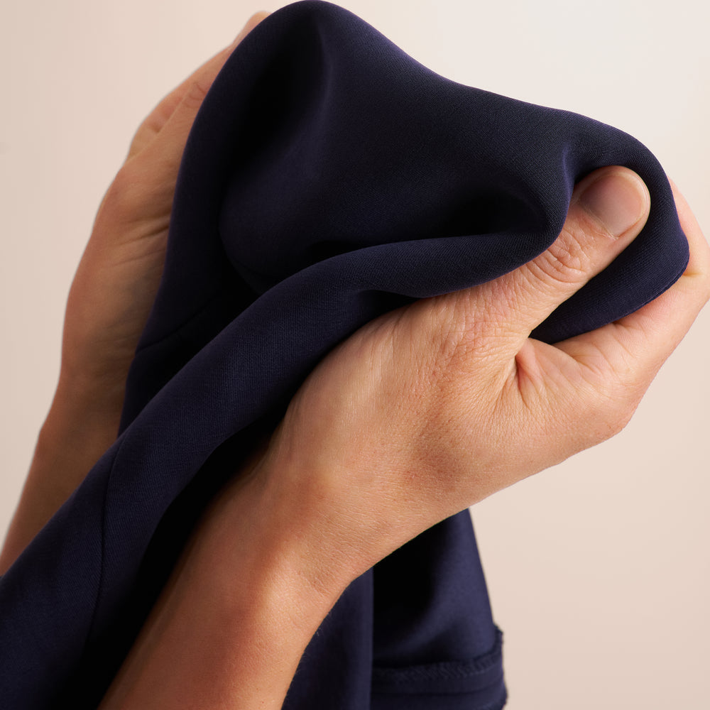 Close-up of a person's hands holding and stretching a piece of dark navy blue fabric against a plain light beige background.
