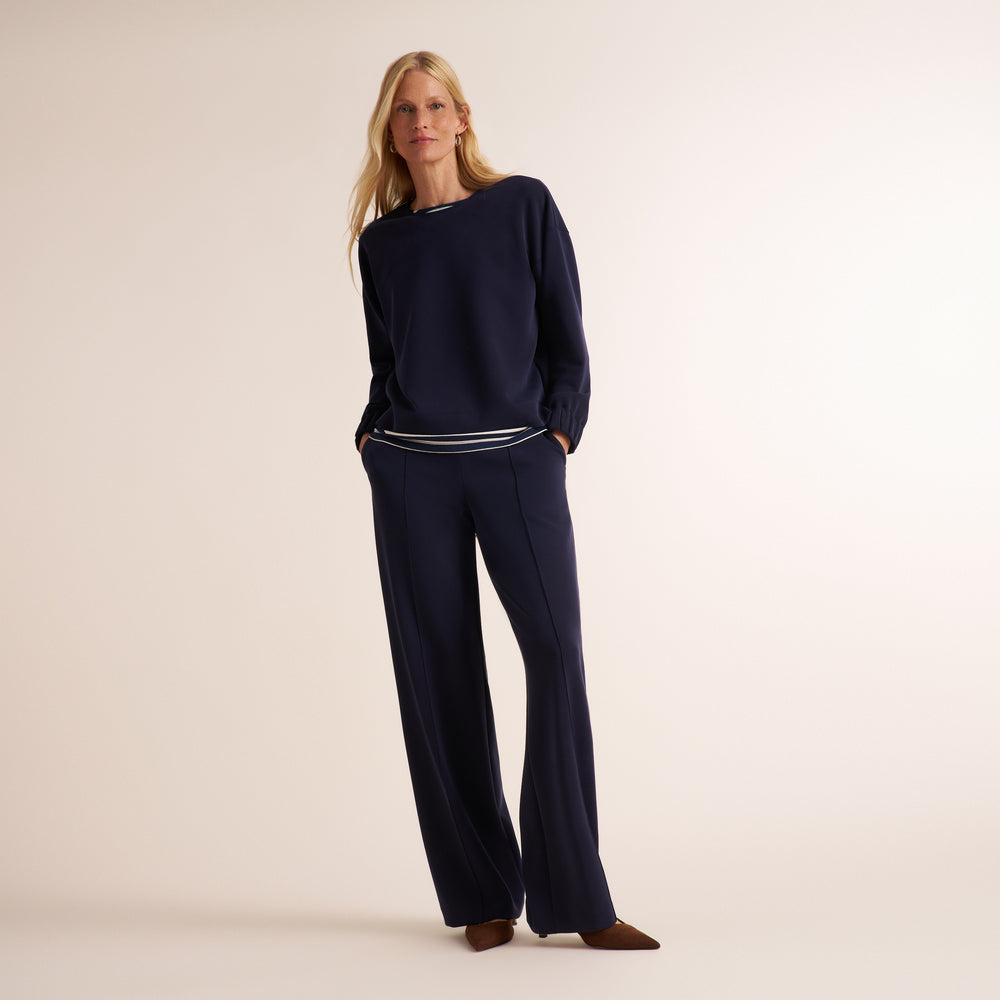 A woman modeling a navy blue knit lounge set consisting of a long-sleeve top and matching wide-leg trousers, styled with brown pointed-toe flats against a warm beige studio background.