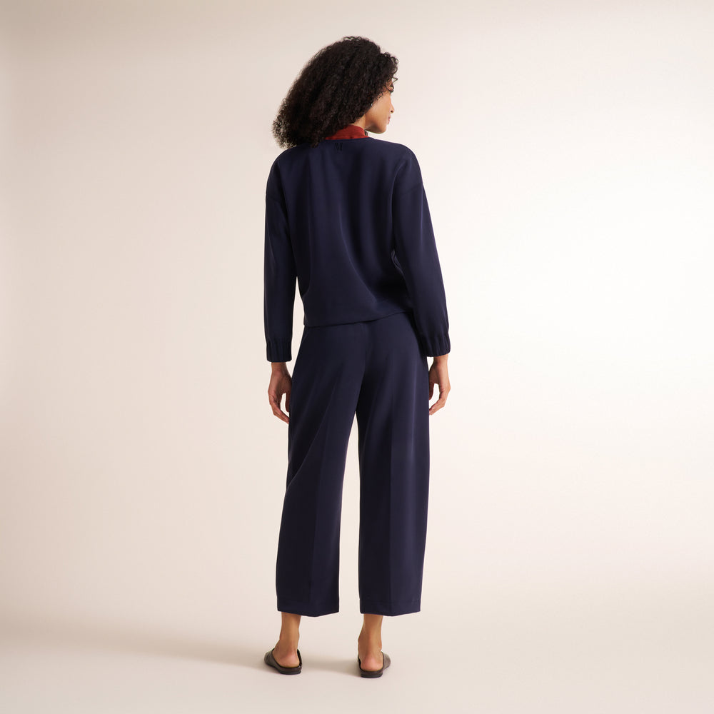 A woman is shown from behind wearing a navy blue long-sleeve top and matching wide-leg cropped pants, photographed against a plain light beige studio background.