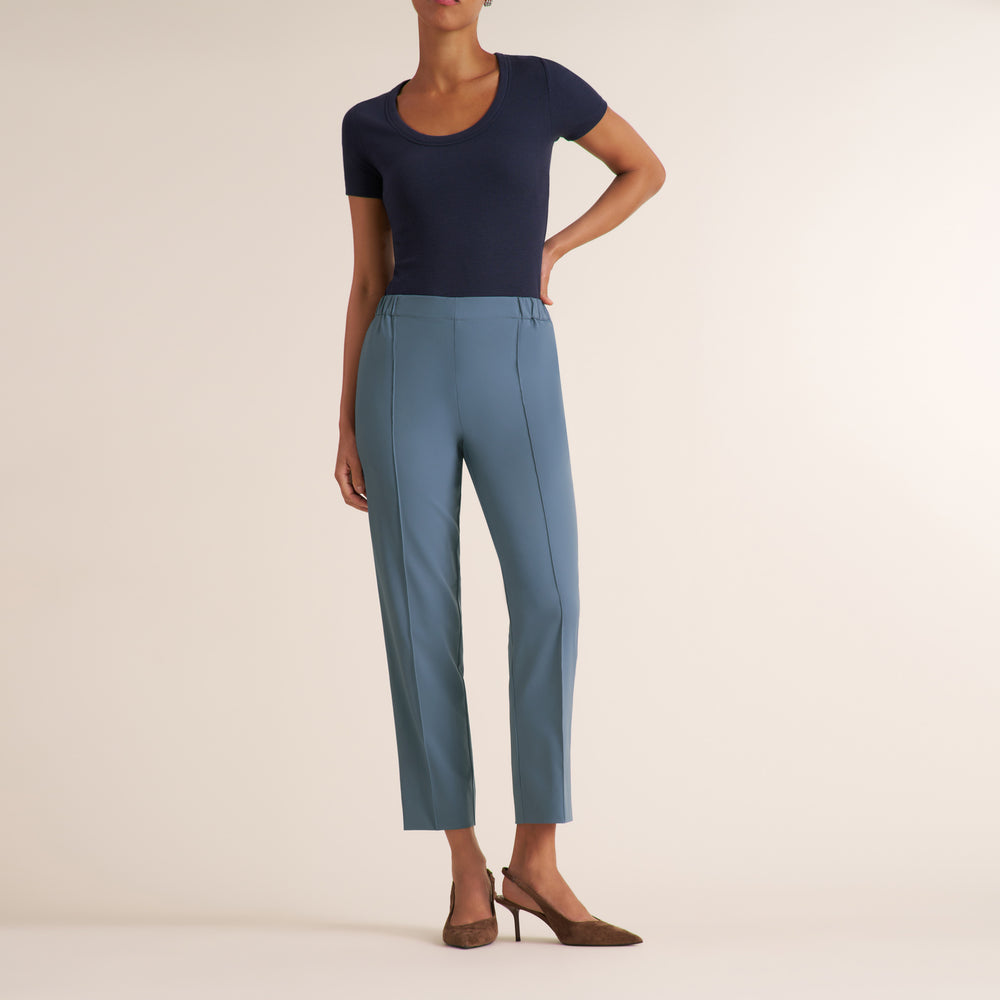 A model wearing slate blue cropped trousers, a navy blue short-sleeve top, and brown pointed-toe heels, standing against a solid light beige studio background.