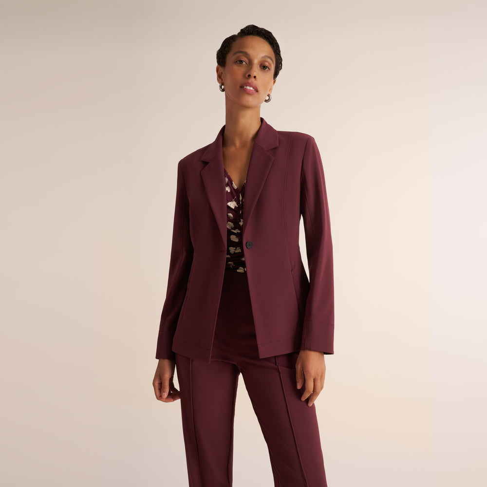 A woman models a matching burgundy two-piece suit consisting of a tailored blazer and straight-leg cropped trousers, paired with a patterned top and tan heels, against a solid light beige studio background.