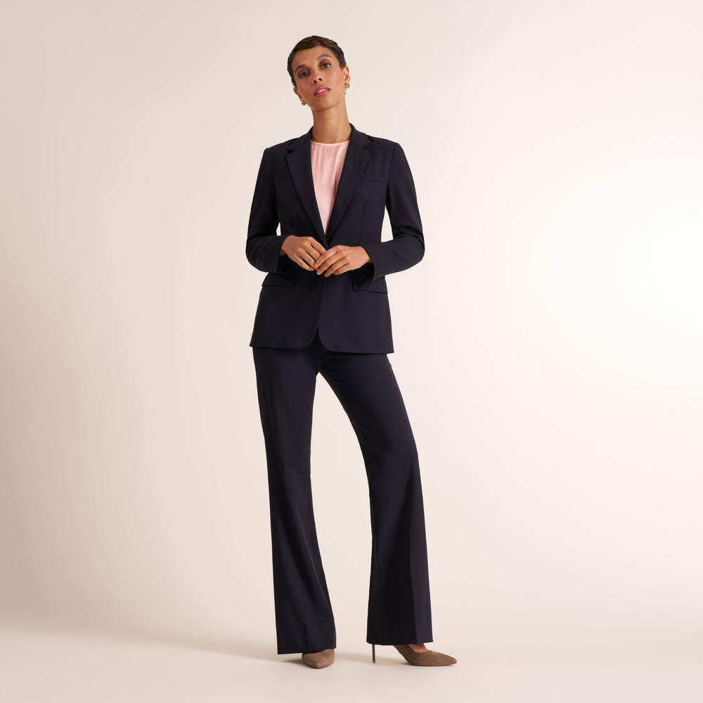A model stands in a studio wearing a dark navy blue two-piece suit consisting of a blazer and matching wide-leg trousers, layered over a light pink top and styled with brown pointed-toe heels.