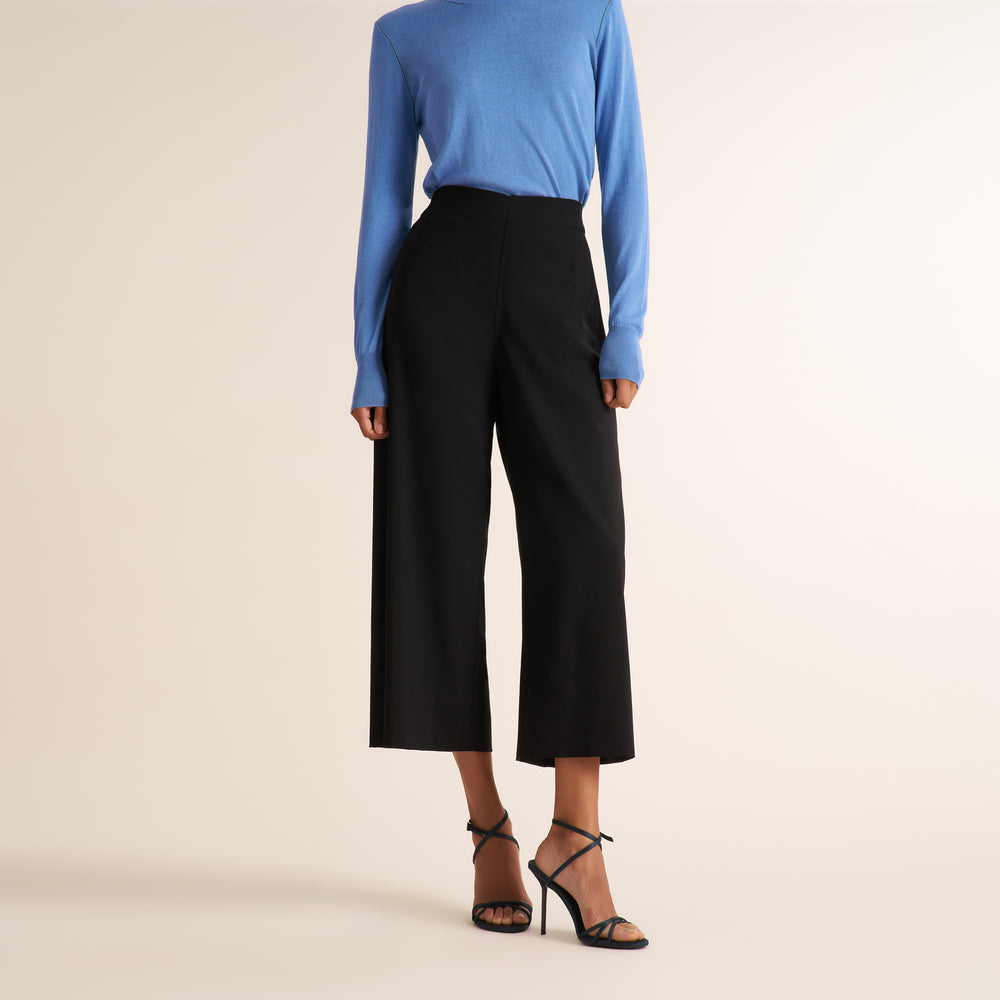 A model shown wearing black wide-leg cropped pants, a blue long-sleeve top, and black high-heeled sandals against a plain beige studio background.