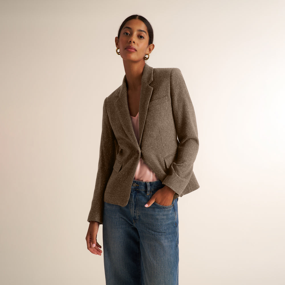 A woman modeling a tailored taupe-brown blazer over a light pink top and blue jeans, standing against a plain warm off-white studio background.