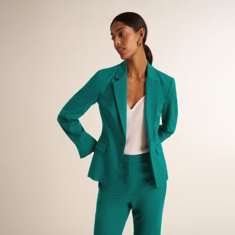 A woman modeling a teal green tailored blazer and matching trousers with a white top, photographed against a plain light beige studio background.