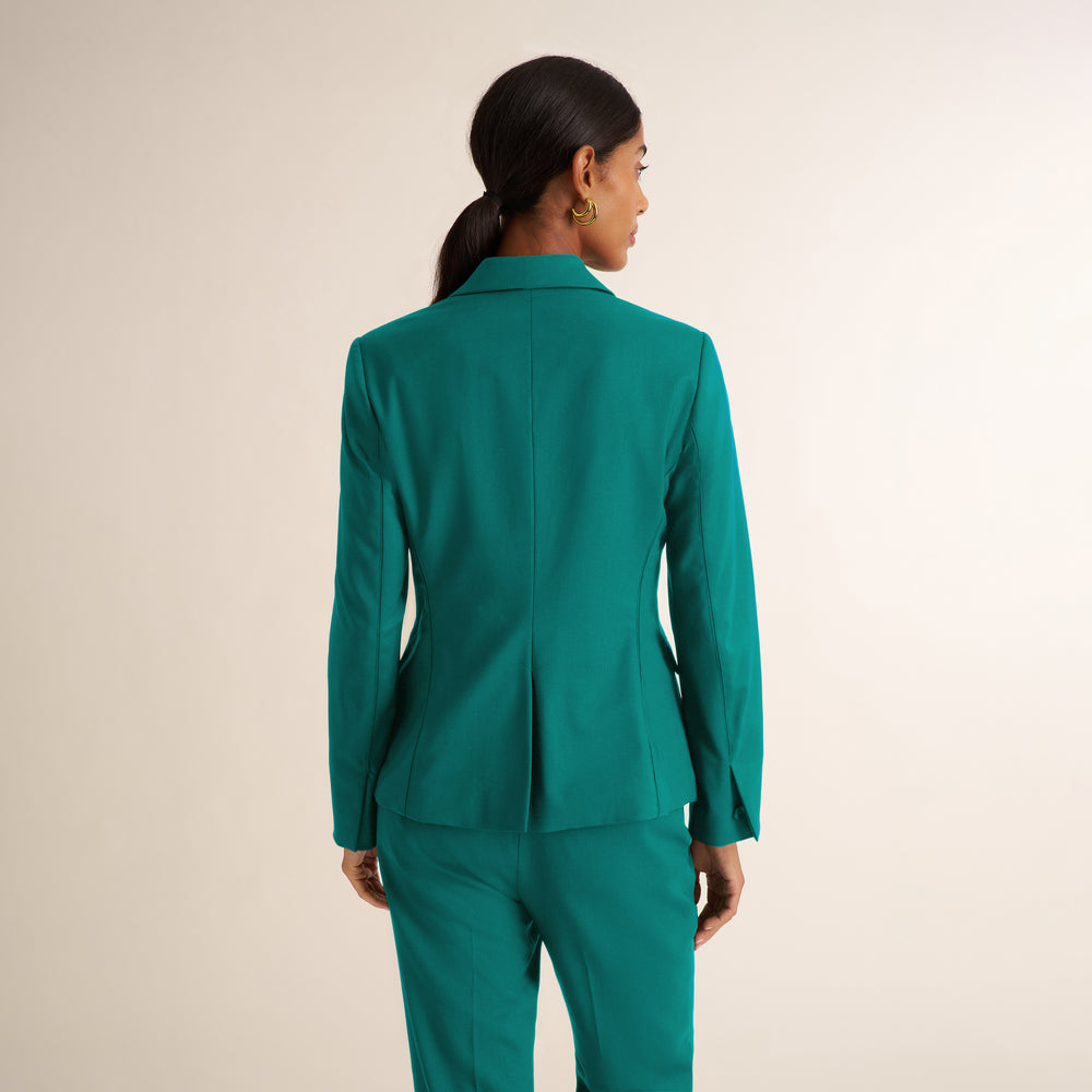 A woman shown from the back wearing a tailored teal-green blazer and matching trousers against a plain light beige studio background.