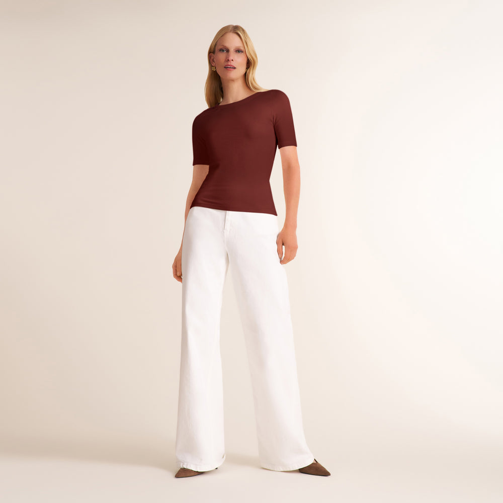 A woman modeling a short-sleeve, asymmetrical neckline top in deep burgundy paired with white wide-leg trousers, standing against a plain beige studio background.
