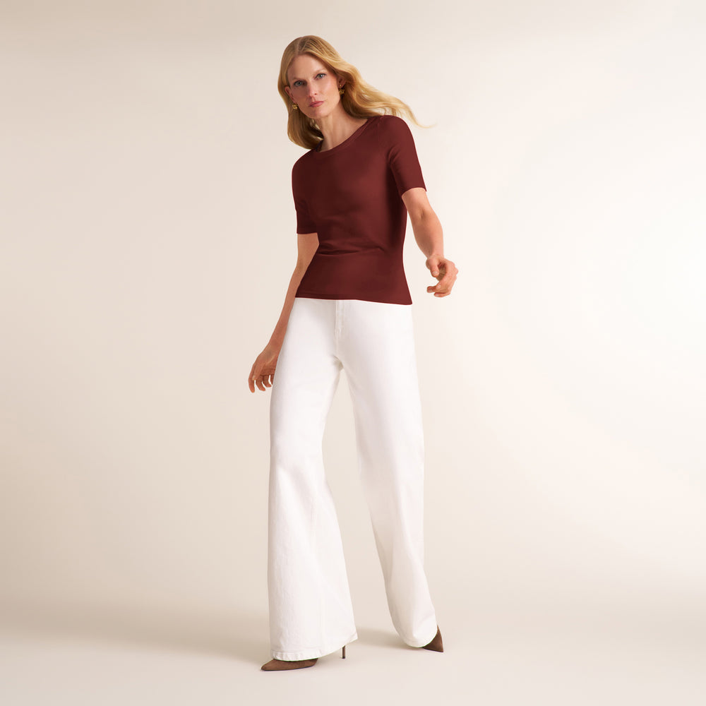 A woman modeling a short-sleeve V-neck knit top in deep burgundy paired with wide-leg white trousers, standing against a plain warm beige studio background.