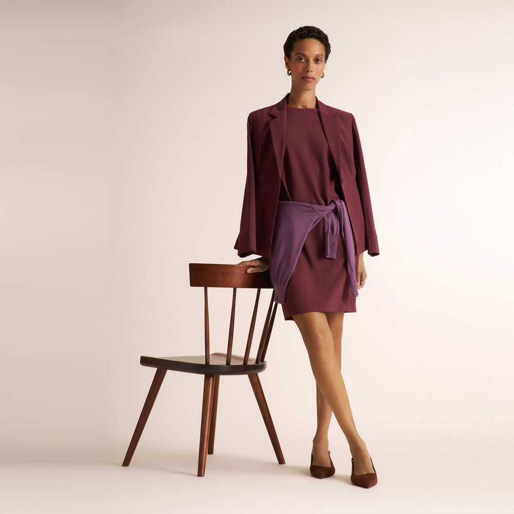 A model stands in a studio wearing a tailored outfit consisting of a dark plum blazer and matching wrap-style mini skirt tied at the waist, paired with brown pointed shoes. She is posed next to a wooden chair against a plain warm off-white background.