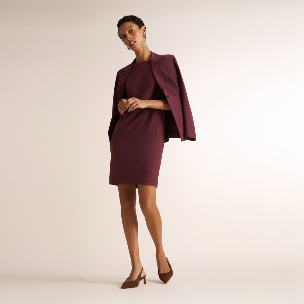 A model poses in a studio wearing a matching burgundy dress and cape-style blazer, paired with brown pointed-toe heels against a warm beige background.
