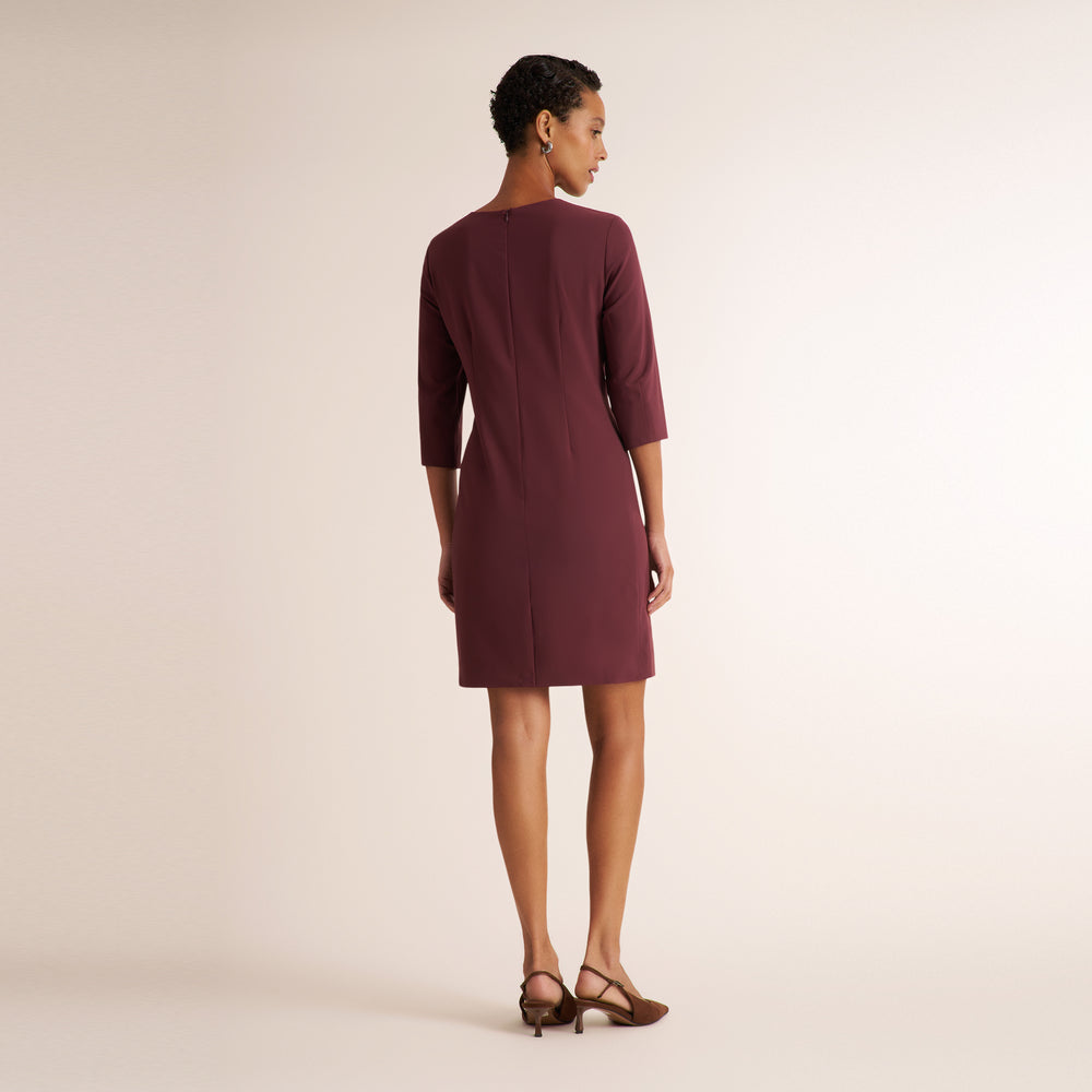 A woman shown from the back wearing a knee-length burgundy dress with three-quarter sleeves and brown strappy heels, standing against a plain light beige studio background.